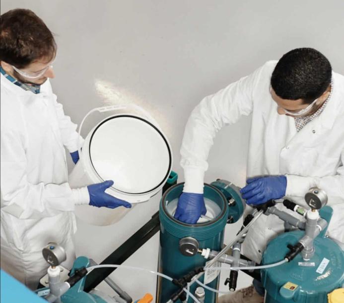Two men in lab coats and safety glasses working in a lab next to two green canisters