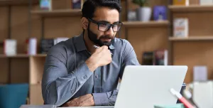 A man with a beard and glasses sitting at a desk looking at a laptop