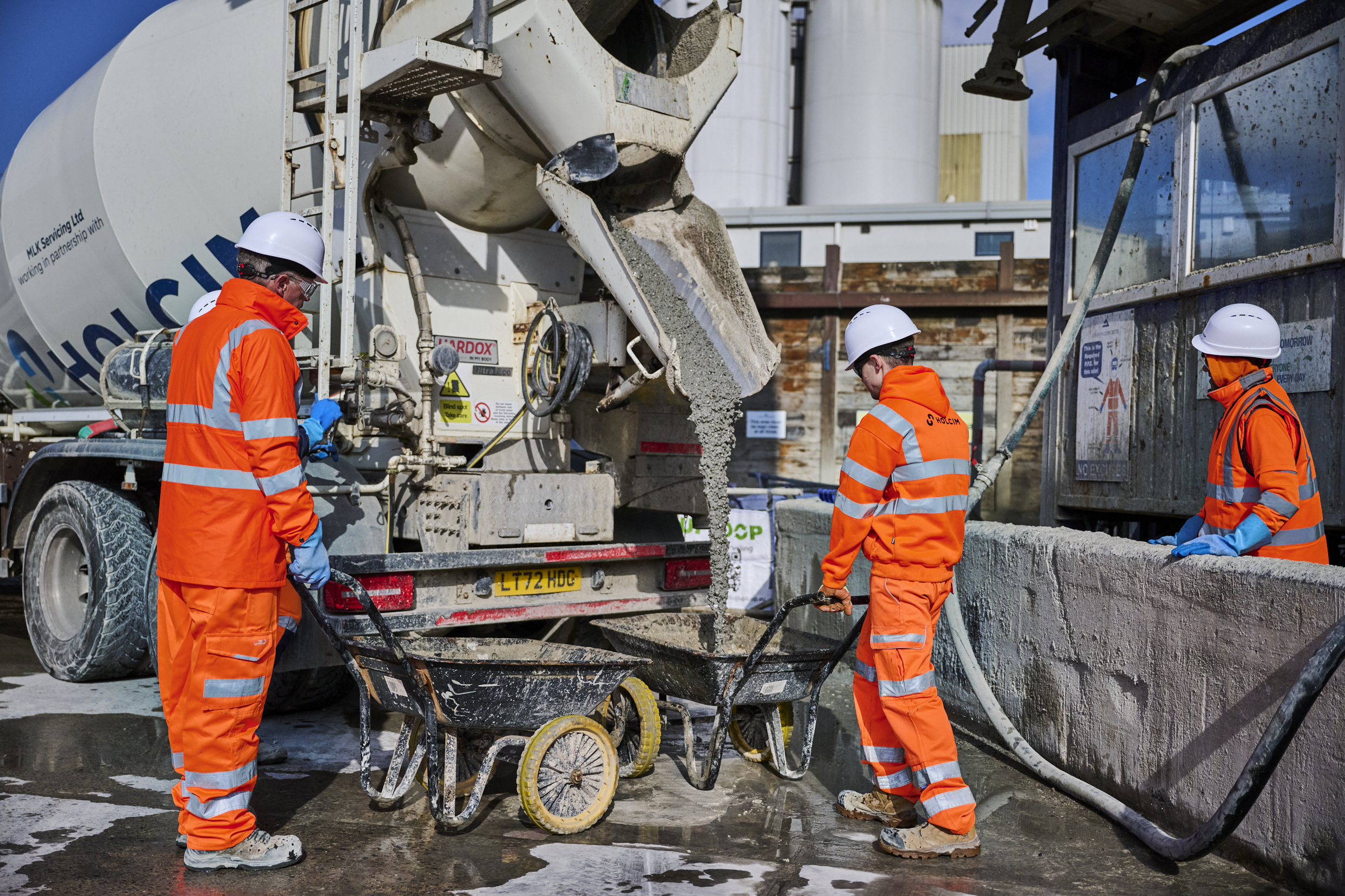 Three men in orange PPE unload cement from a Holcim branded cement truck