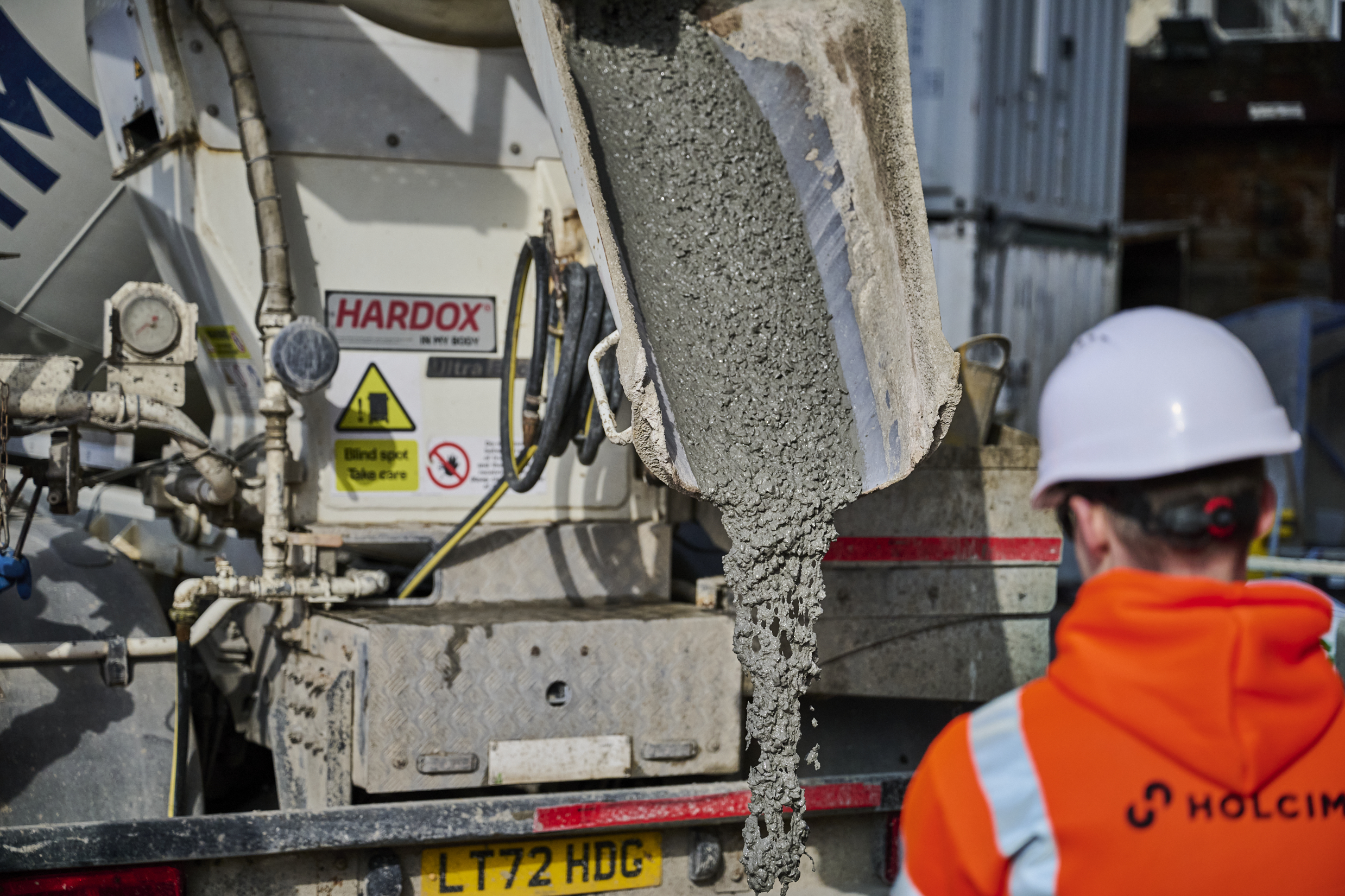 A close up image of cement being unloaded from a Holcim cement truck watched by an operative in orange PPE and a white hard hat.