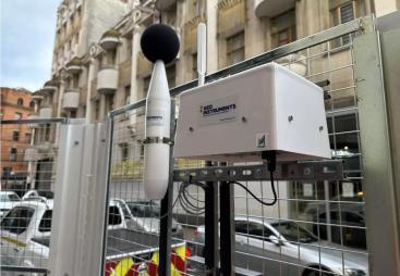 a dust monitoring station attached to a metal fence withan building in the background