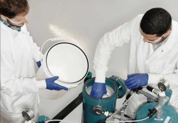 Two men in lab coats and safety glasses working in a lab next to two green canisters