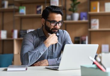 A man with a beard and glasses sitting at a desk looking at a laptop
