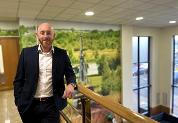 A bald man with a beard in an open necked shirt and dark suit standing on an internal office landing