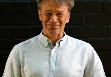 A head and shoulders picture of a man with unruly hair in a white shirt against a black background