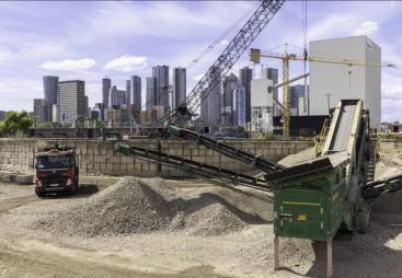Piles of aggregate in a yard with a truck and green screen with a crane and tall buildings in the background