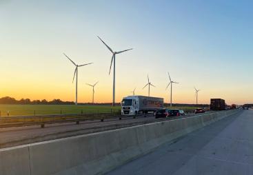 A truck on a motorway with a concrete safety barrier in the foreground and several wind turbines in the background.