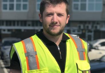 A man with short dark hair wearing yellow PPE in front of a mid-rise building