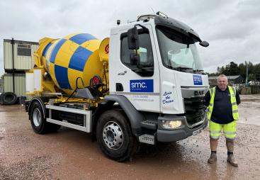 A man in yellow PPE stands in front of a white truck fitted with a yellow and blue concrete mixer