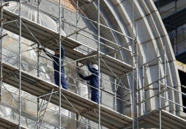 Two men in blue overalls and white hard hats on a scaffold cleaning a building