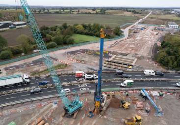 A green crane and yellow piling rig photographed from the air installing piling on a large construction site by a dual carriageway