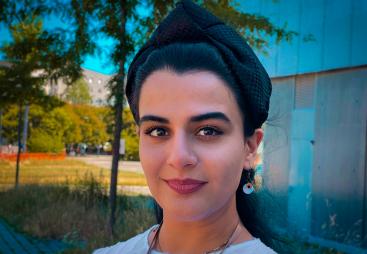 Head and shoulders picture of a young woman with dark hair, white jumper and necklace outside with trees in the background