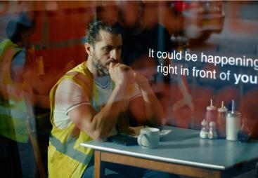 A man with dark hair in a top knot, wearing yellow PPE sitting at a cafe table looking anxious