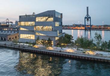 A seven storey concrete building next to a body of water with a ship loading crane in the background