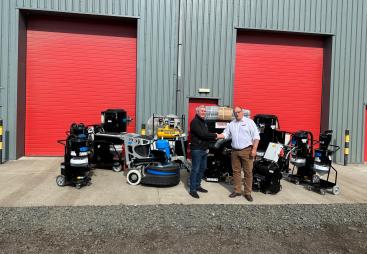 Two men shake hands infront of a range of floor grinding equipment, which is placed outside a grey warehouse with two, big red doors
