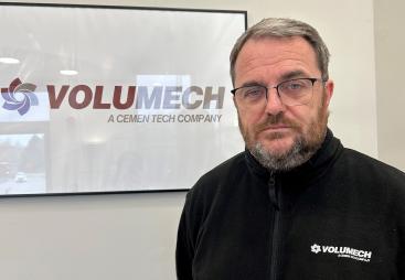A man with grey hair, beard and glasses in a black fleece in front of a sign on the wall in an office
