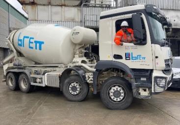 A white concrete mixer truck being driven by a man in red PPE