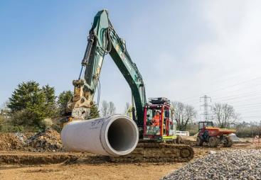 A green excavator with yellow pipe-handling grab moves a large piece of concrete pipe