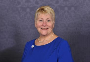A head and shoulders photo of Caroline Gumble, CEO, Chartered Institute of Building (CIOB), wearing a blue dress, brooch and necklace.