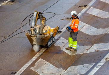 A man in orange, grey and yellow PPE uses a remote controlled piece of construction equipment fitted with a breaker to carry out repairs to a concrete road