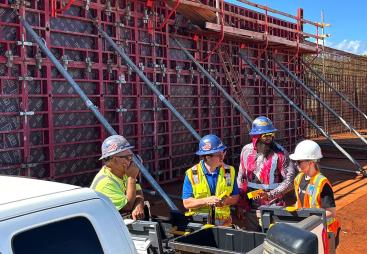 A group of construction workers in various colours of PPE stand in front of a wall of formwork