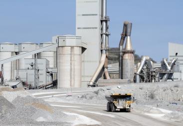 A yellow Caterpiller rigid dump truck (RDT) drives away from a huge cement factory 