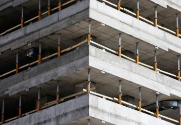 This image depicts a close-up view of a concrete multi-story building under construction, highlighting the raw structural elements such as concrete slabs and metal supports. The image conveys a sense of industrial progress and urban development in a modern city.