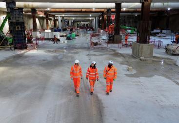 Three people in orange PPE walk across the competed base slab in the 850m long underground station box at Old Oak Common.