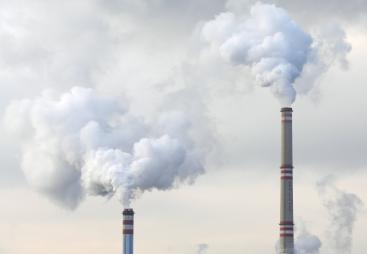 The top of two tall chimneys against a grey sky billow "smoke" into the atmosphere