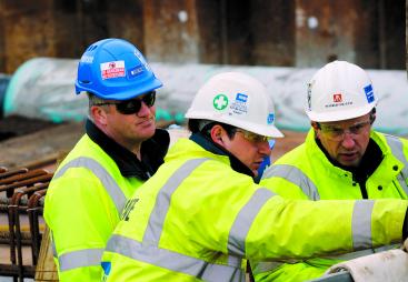 Three Costain construction workers in yellow PPE look at plans on a construction site.