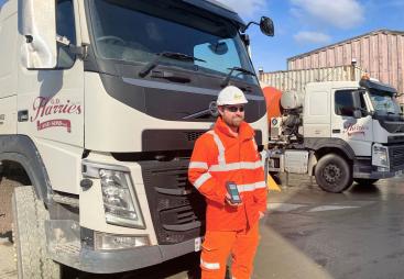 A man in orange PPE standing in front of a cement mixer holding a mobile device that uses the Podfather app to access their schedules and delivery notes, navigate routes, and record delivery details digitally. 