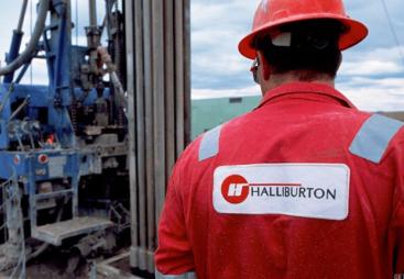 A man in red overalls and red hard hat stands with his back to the camera, a Halliburton logo on his back, surveying a Halliburton Energy Services project.