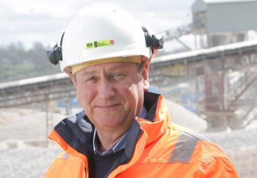 A head and shoulders picture of Mike Phillips, CEO of the British Aggregates Association, wearing orange PPE and a white hard hat, in a quarry.