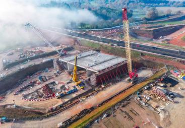 An aerial view of the A46 Kenilworth Bypass box structure under construction with a large yellow and red crawler crane and a smaller white telescopic crane and white crawler crane. Image: HS2.