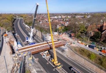 An aerial view of the new M67 St Annes Bridge under construction with a yellow crane on the near side of the bridge and a white crane on the far side.