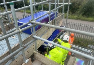 Two people in yellow PPE carry out a bridge inspection from a scaffold suspended from the side of the bridge