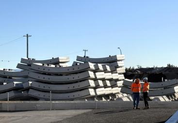 Two men in orange PPE stand next to a stack of precast concrete tunnel segments at the Western Harbour Tunnel Precasting facility in Emu Plains.