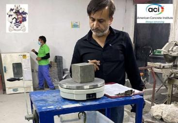 A man with a beard, waring a dark shirt inspects a concrete cube in a testing laboratory.