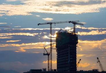 A high rise building framed against a sunset flanked by several tower cranes