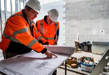 Two men in orange PPE look at plans on a table on a construction site
