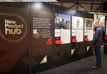 A man stands right of frame looking at entries to the UK Concrete Show's New Product Hub, which are displayed on a large, floor standing board.