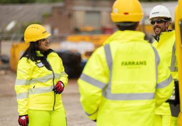 Three people in yellow PPE stand talking with construction machinery in the background