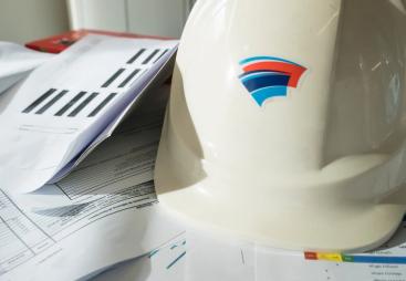 A white hard hat with red and blue insignia, and technical construction papers on a table