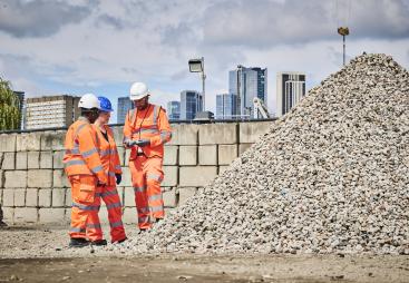 Three people in orange PPE stand discussing aggregate needs in a construction materials yard with a wall and city scape in the background