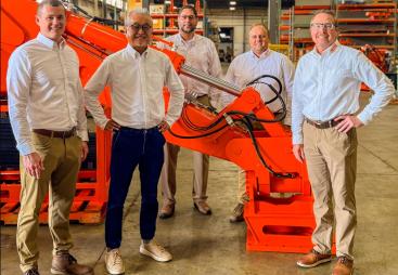 Five men dressed in smart shirts and trousers stand around a large red excavator arm