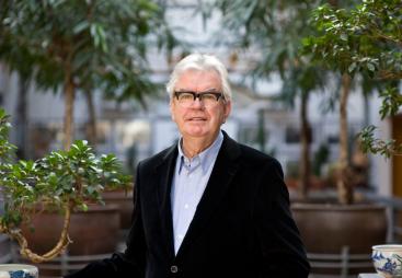 A grey-haired Sir Terry Farrell (1938-2025) wearing glasses, a dark jacket and light blue shirt photographed in front of an avenue of small trees in an urban setting © richard gleed / farrell