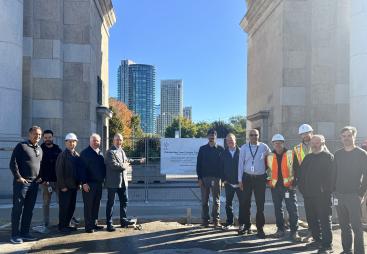 A group of people in business wear and PPE celebrate the use of Bio Graphene Solutions’ graphene-enhanced low-carbon concrete beneath Toronto’s historic Princes’ Gates at Exhibition Place.