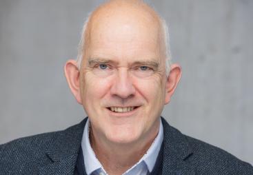 Head and shoulders photo of a bald, smiling man in open necked shirt, jumper and jacket