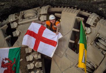 A man wearing a hard hat and waving a Cross of St George flag pokes his head through a TBM cutter wheel with a Welsh flag and another unidentified flag adorning the cutter head as HS2 engineers celebrate the breakthrough of tunnel boring machine Elizabeth at Washwood Heath in Birmingham, completing the 3.5-mile Bromford tunnel under the M6. (Image © HS2 Ltd)
