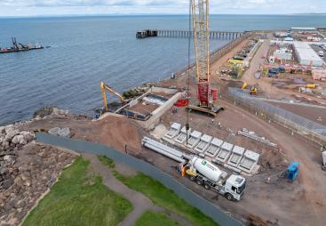A red and yellow crawler crane lifts precast concrete elements at a shoreline construction site
